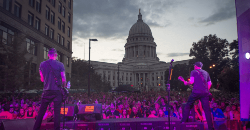 A band performs on stage in front of a large crowd, with a historic building and dome in the background at sunset.