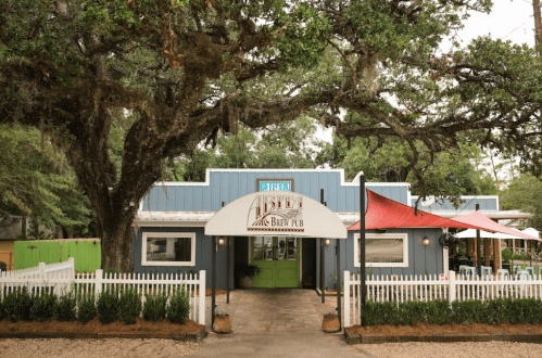 A charming brew pub with a white awning, surrounded by trees and greenery, featuring outdoor seating.
