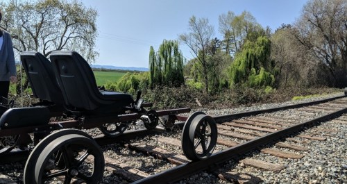 A two-seat rail cart on a railway track, surrounded by greenery and trees under a clear blue sky.