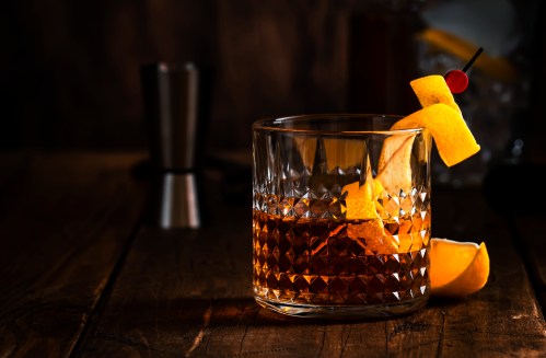 A glass of whiskey with a twist of lemon and a cherry, set on a wooden table with a dimly lit background.
