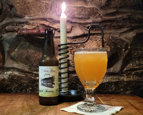A bottle of beer, a glass of orange drink, and a candle on a rustic wooden table against a stone wall.