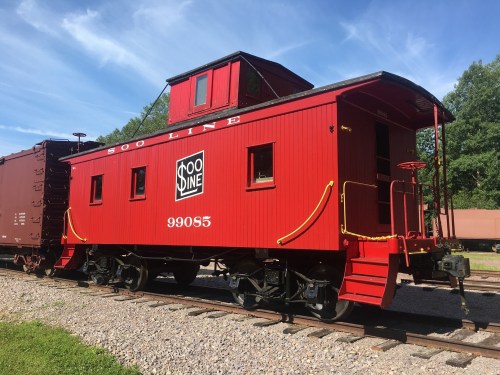 A bright red caboose labeled "Soo Line 99085" parked on a gravel track under a blue sky.