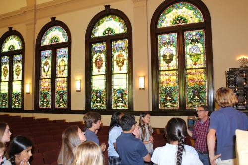 A group of people gathers indoors, admiring colorful stained glass windows in a spacious room.