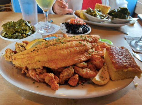 A plate of fried fish and shrimp, served with cornbread, black beans, collard greens, and lemon wedges.