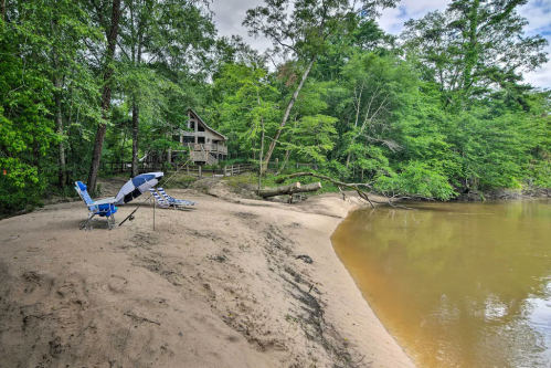 A sandy riverbank with lounge chairs and an umbrella, surrounded by lush greenery and a cabin in the background.