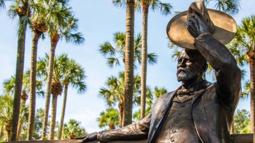 A bronze statue of a man tipping his hat, surrounded by palm trees under a clear blue sky.