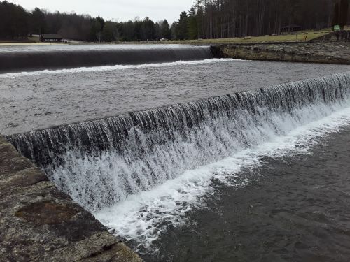 A serene view of a waterfall cascading over a stone dam, surrounded by trees and a calm river.