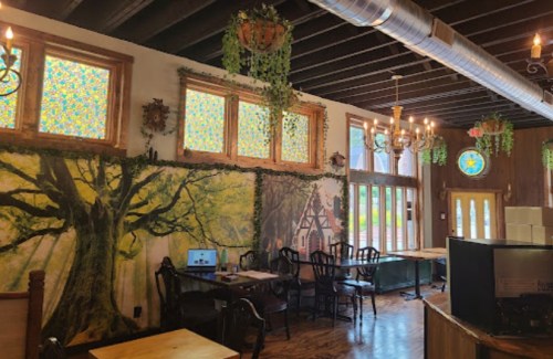 Cozy restaurant interior with stained glass windows, tree mural, wooden tables, and hanging plants.
