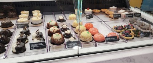 A display case filled with various desserts, including cakes, pastries, and tarts, each labeled with names and prices.