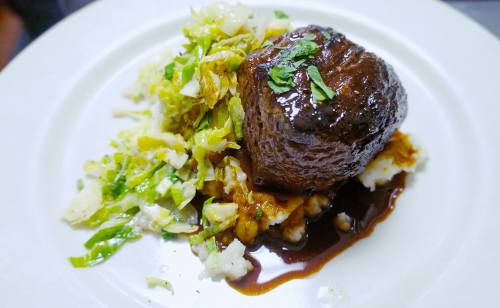A plated dish featuring a tender beef filet on a bed of mashed potatoes, topped with shredded greens and a rich sauce.