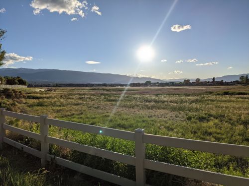A sunlit landscape with a grassy field, mountains in the background, and a white fence in the foreground.