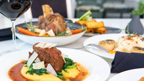 A plate of braised beef on polenta with greens, garnished with cheese, alongside wine and other dishes in the background.