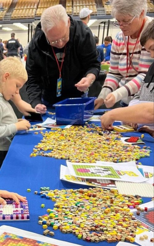 A group of people, including a child, work together at a table covered in colorful tiles and game materials.