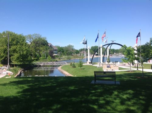 A scenic park by a lake featuring flags, a gazebo, and green lawns under a clear blue sky.
