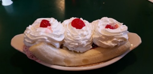 A banana split topped with three dollops of whipped cream and cherries, served in a dish on a green table.