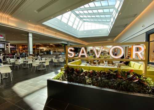 A bright food court with a "SAVOR" sign, featuring seating areas and large windows letting in natural light.