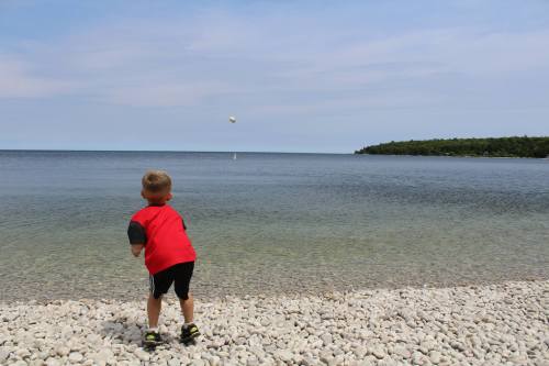 A young boy in a red shirt throws a stone into a calm lake, with a rocky shore and trees in the background.