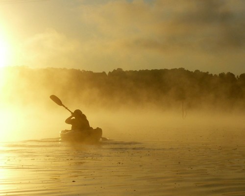 A person kayaking through a misty lake at sunrise, with golden light illuminating the scene.