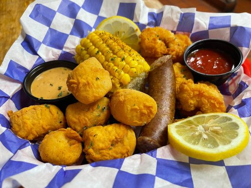 A basket of fried seafood, corn on the cob, sausage, and lemon slices, served with dipping sauces on a checkered paper.