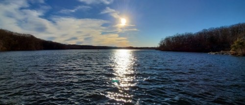 A serene lake scene with sunlight reflecting on the water, surrounded by trees and a clear blue sky.