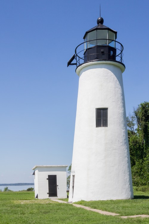 A white lighthouse stands beside a small building, set against a clear blue sky and green grass.