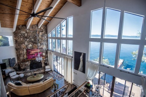 Spacious living room with large windows overlooking a lake, featuring a stone fireplace and modern decor.