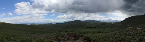 A panoramic view of rolling green hills under a cloudy sky, with distant mountains and a winding road.