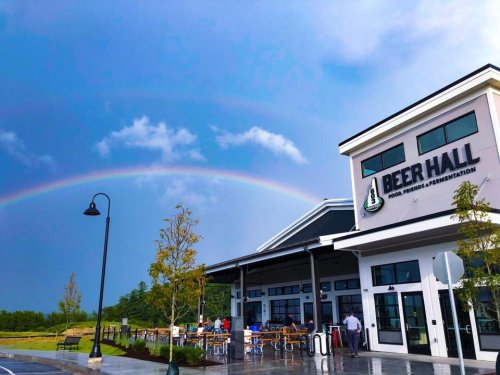 A modern beer hall with a rainbow in the sky, surrounded by greenery and people enjoying the outdoor seating.
