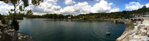 A panoramic view of a serene lake surrounded by trees and rocky shores, with people enjoying the water on a sunny day.