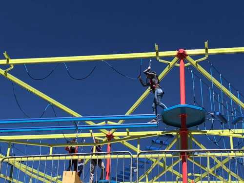 Two people navigate a colorful ropes course against a clear blue sky.