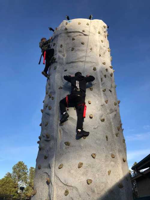 Two climbers ascend a tall rock wall with grips, set against a clear blue sky.