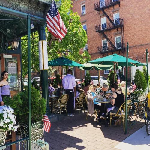 Outdoor café scene with diners under green umbrellas, American flags, and a brick building in the background.