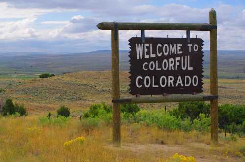 A wooden sign reading "Welcome to Colorful Colorado" stands against a backdrop of rolling hills and cloudy skies.