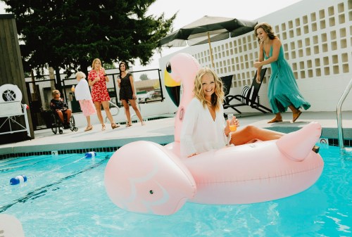 A woman relaxes on a pink flamingo float in a pool, while friends enjoy the sun and laughter nearby.