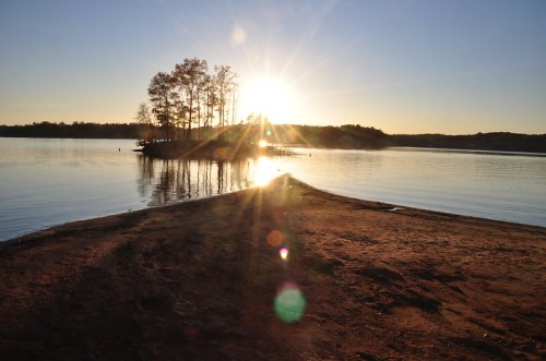 Sunset over a calm lake, with an island and trees silhouetted against the glowing horizon.