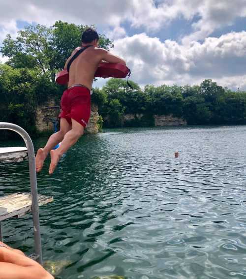 A person jumps off a diving board into a clear lake surrounded by trees and rocky cliffs.