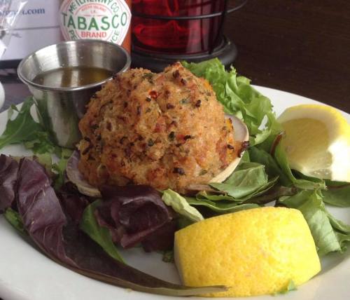 A plate featuring a crab cake on greens, a lemon wedge, and a small cup of sauce, with a bottle of Tabasco in the background.