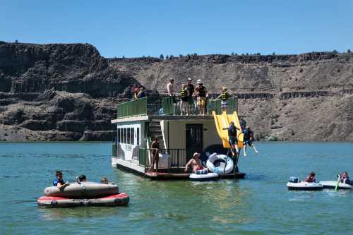 A houseboat on a lake with people swimming, sliding, and relaxing on inflatable rafts under a clear blue sky.