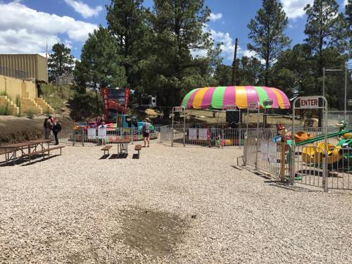 A colorful amusement area with rides, picnic tables, and trees under a blue sky. Fenced entrance visible.