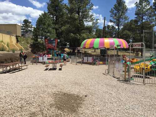 A colorful amusement area with rides, picnic tables, and trees under a blue sky. Fenced entrance visible.