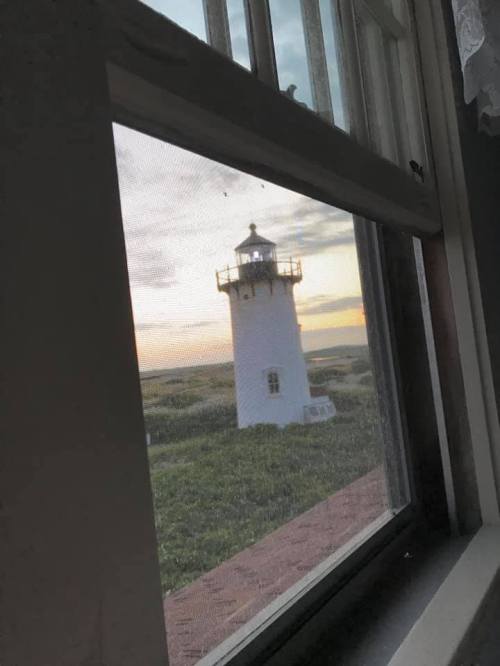 View of a white lighthouse through a window, with a sunset sky and greenery in the background.
