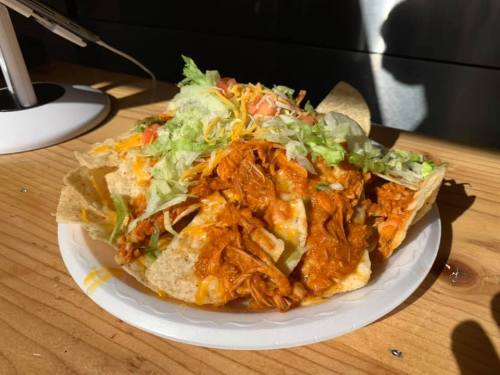 A plate of nachos topped with shredded chicken, lettuce, tomatoes, and cheese on a wooden table.