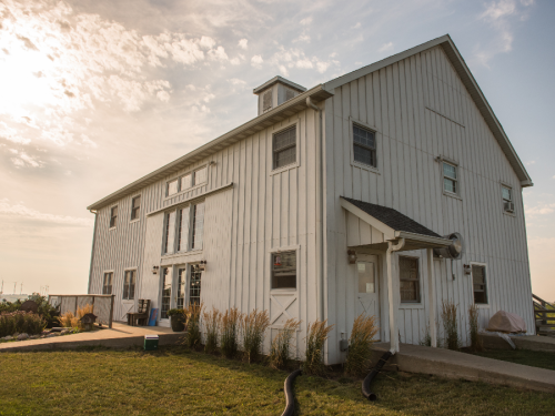 A white barn-style building with large windows, surrounded by grass and plants, under a partly cloudy sky.