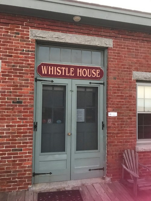 A brick building with a green door labeled "Whistle House" and a wooden chair nearby.