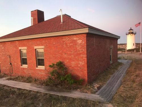 A red brick building with a wooden walkway, near a lighthouse and an American flag in the background.