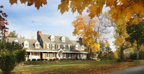 A large, charming house surrounded by colorful autumn trees and a well-maintained lawn under a clear blue sky.