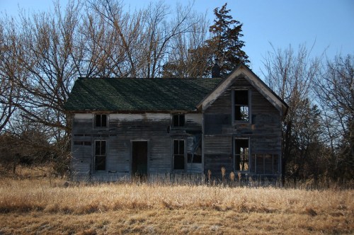 Abandoned wooden house with broken windows, surrounded by tall grass and bare trees under a clear blue sky.