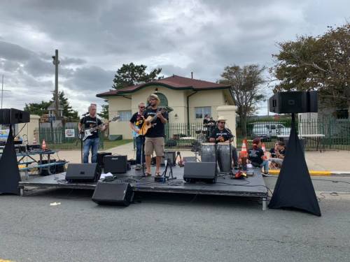 A band performs on an outdoor stage with instruments, while people sit nearby on the ground. Overcast sky in the background.