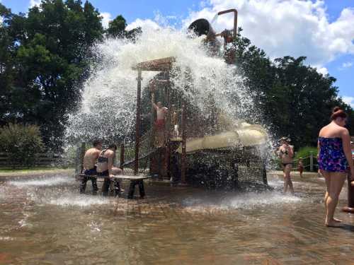 Children playing in a splash pad with a water slide, surrounded by splashing water and sunny skies.