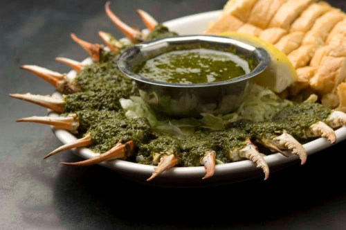 A plate of crab claws arranged around a bowl of green sauce, with lemon slices and bread rolls on the side.